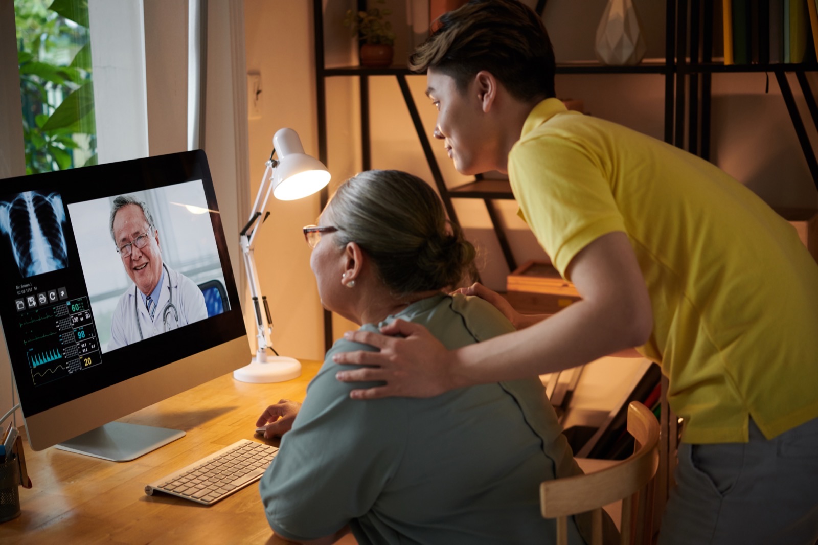 A son with his hand on his mother's shoulder as they join a video consultation with a physician together